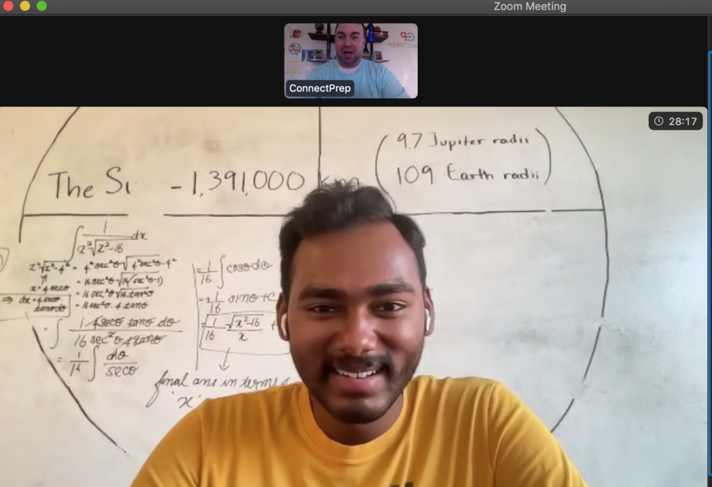 A young man in a yellow shirt smiles during a Zoom meeting. Behind him is a whiteboard with math equations and notes mentioning Jupiter radii and Earth radii. Another person appears in a small window at the top.