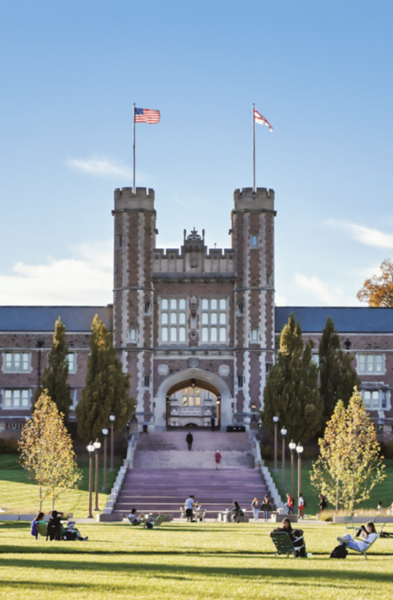 A large gothic-style university building with two towers and arched entrance, American flags atop each tower, students relaxing and walking on the grassy lawn under a clear blue sky.