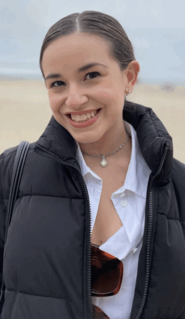 A young woman with dark hair tied back smiles at the camera. She is wearing a black puffy jacket over a white shirt, a necklace, and has sunglasses hanging from her shirt. The background is a sandy outdoor area.