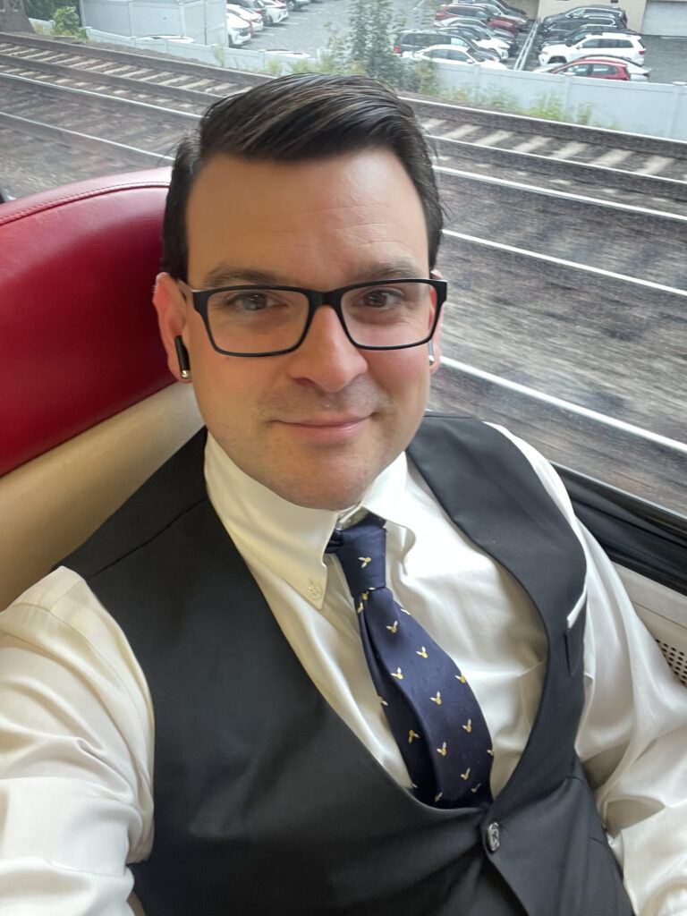 A man wearing glasses, a white shirt, a patterned tie, and a dark vest sits by a train window. He is smiling slightly, wearing wireless earbuds, and train tracks and parked cars are visible outside the window.