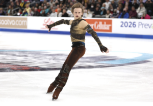 A figure skater in a brown costume performs on the ice with one arm extended and one leg bent, in front of a large audience at an indoor arena.